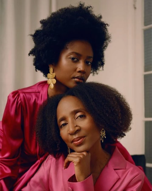 Two African American women in pink satin tops pose indoors. One woman is resting her head on the other's shoulder.