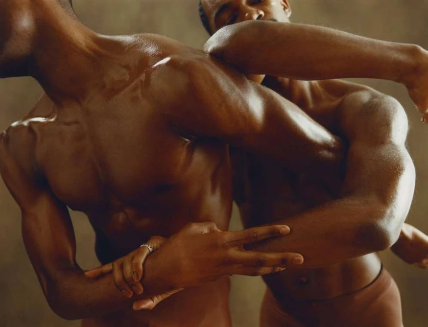 Striking studio shot of two shirtless African American men in close embrace, showcasing muscles and physique. Dark skin tones, neutral background.