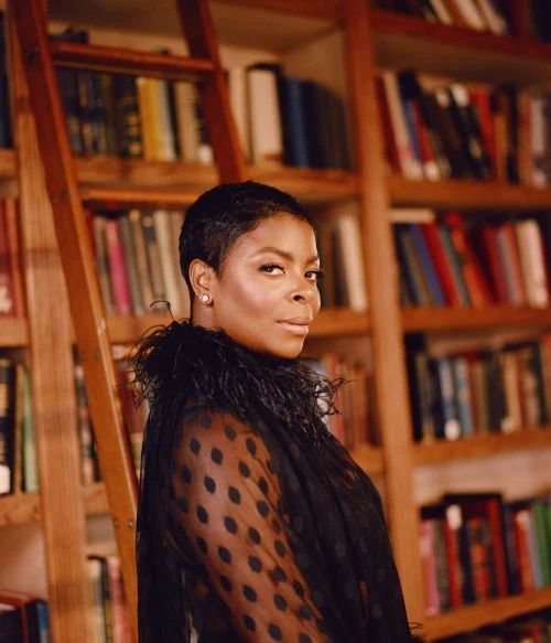 Portrait of an adult woman with a short haircut, wearing a sheer polka dot top with a feathered collar in front of a bookshelf and ladder.