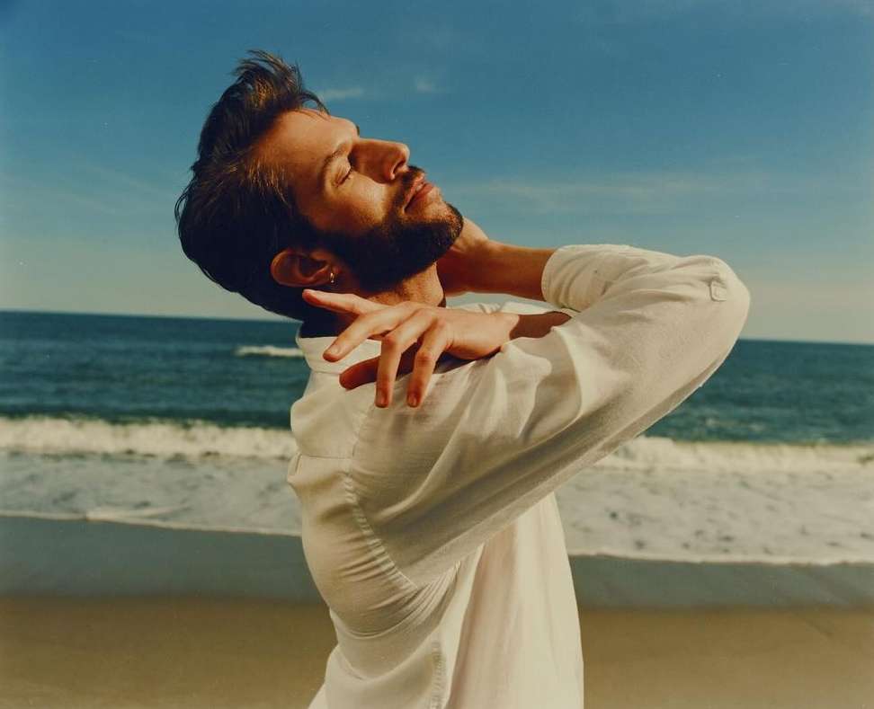 Man with beard wearing a white button-down shirt posing on a sandy beach with the ocean in the background by Kendall Bessent