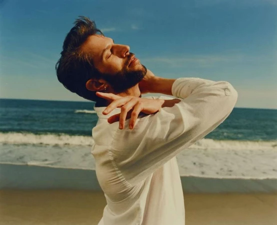 Man with beard wearing a white button-down shirt posing on a sandy beach with the ocean in the background by Kendall Bessent