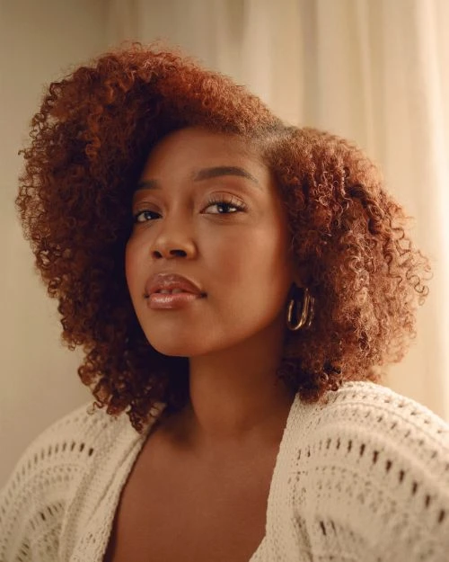 Close-up portrait of a beautiful Black woman with auburn curly hair, wearing gold hoop earrings and a white knitted cardigan.