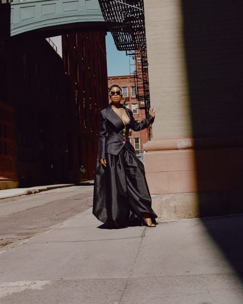 Stylish African-American woman in a dramatic black blazer and maxi skirt poses in an urban setting, accented by sun and shadow.
