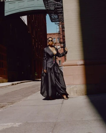 Stylish African-American woman in a dramatic black blazer and maxi skirt poses in an urban setting, accented by sun and shadow.