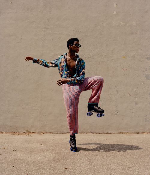 Young man in a colorful patterned shirt and pink pants strikes a pose on roller skates against a neutral urban background in bright daylight.