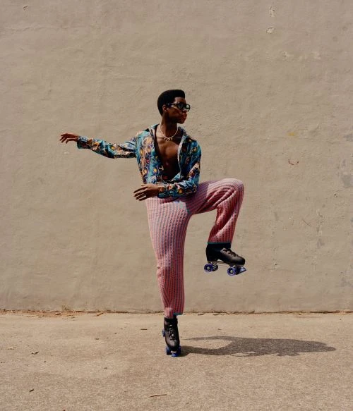 Young man in a colorful patterned shirt and pink pants strikes a pose on roller skates against a neutral urban background in bright daylight.