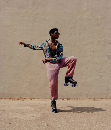 Young man in a colorful patterned shirt and pink pants strikes a pose on roller skates against a neutral urban background in bright daylight.