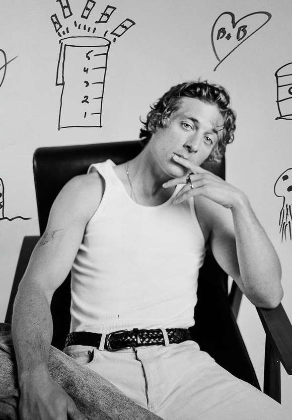 Black and white portrait of a man in a tank top sitting in a chair smoking a cigarette in front of a wall with doodles.