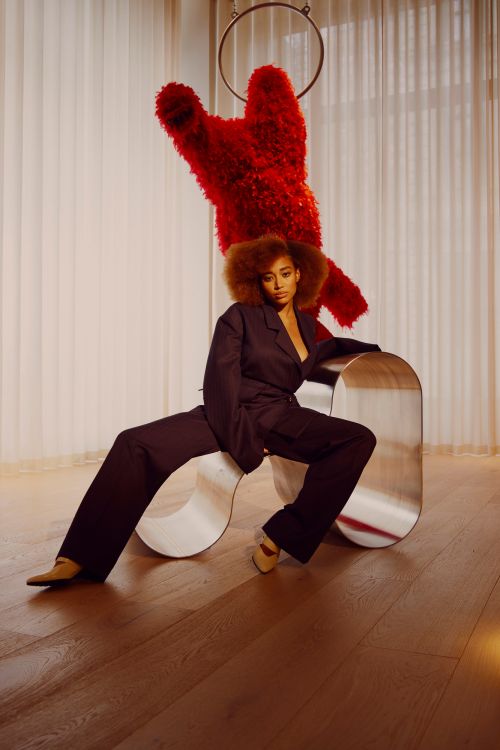 Adult woman in a dark suit poses indoors on a metal bench. Behind her hangs a large red, decorative, bear-like object in a ring.