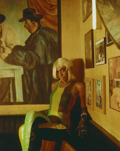 Woman with platinum bob wig poses on a chair wearing green and red tank top in a room with paintings and yellow walls.
