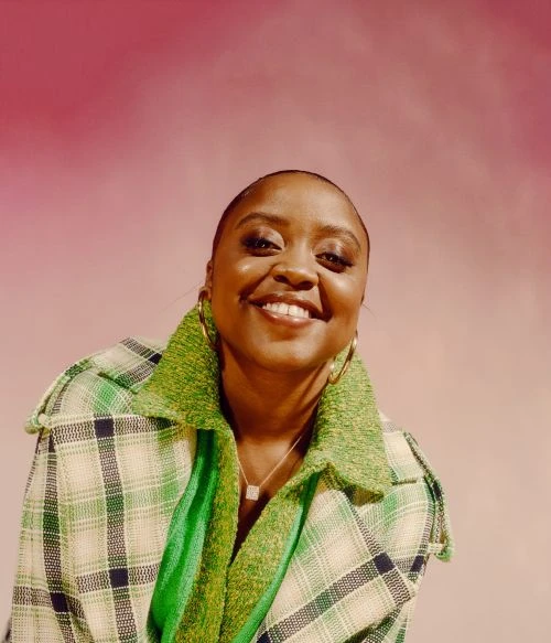 Headshot of smiling adult Black woman in green plaid coat and gold hoop earrings against a pink-toned gradient background.