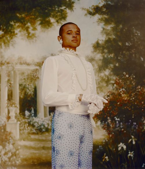 Portrait of a person with short hair, wearing a ruffled white shirt, pearl necklace, and beaded pants, against a floral painted backdrop.