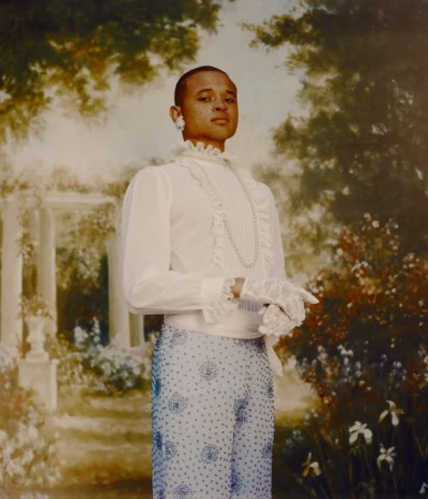 Portrait of a person with short hair, wearing a ruffled white shirt, pearl necklace, and beaded pants, against a floral painted backdrop.