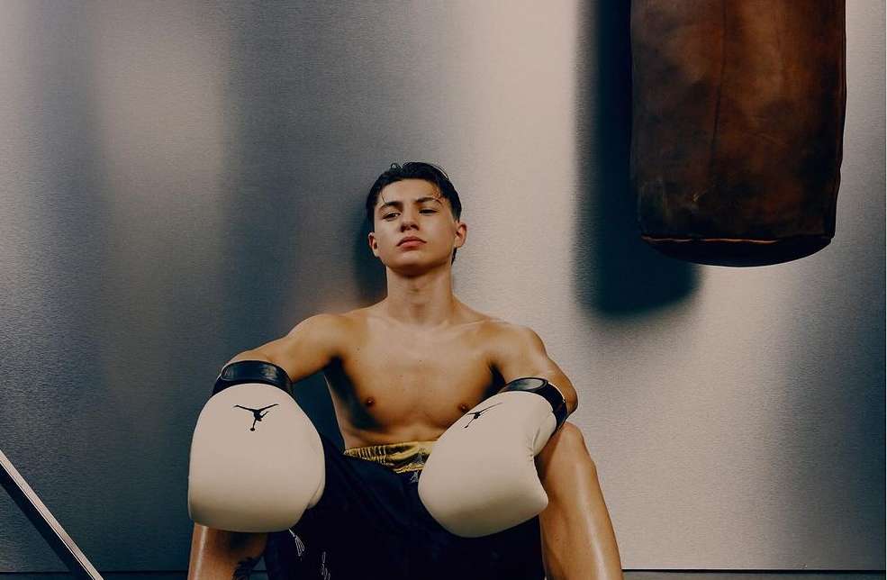 Shirtless male boxer with boxing gloves sits against a gray wall in workout gear next to a punching bag, bathed in studio light.