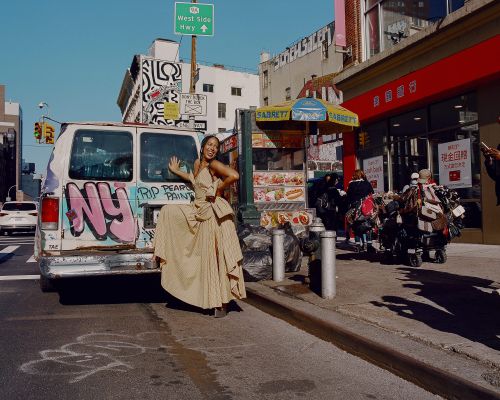Woman in elegant dress poses in front of graffiti-covered van on a city street near Sabrett food cart under a bright blue sky.