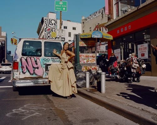 Woman in elegant dress poses in front of graffiti-covered van on a city street near Sabrett food cart under a bright blue sky.