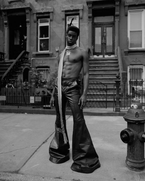 Black and white portrait of a man in bell-bottoms posing outdoors in front of a brick building with a ribbon and bare chest.