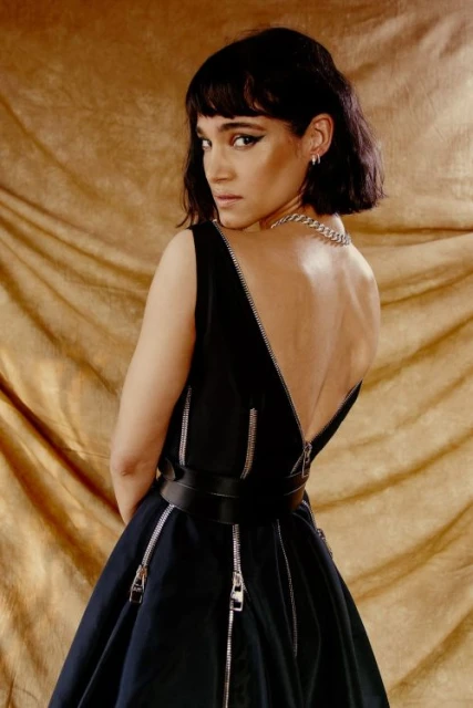 Brunette woman in a black dress with zipper details poses against a textured gold background, looking over her shoulder.