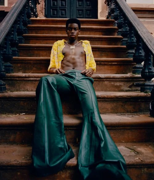 A young black man sits on outdoor steps wearing green bell-bottoms, a yellow crochet shirt, and a chain; outdoor fashion shot.
