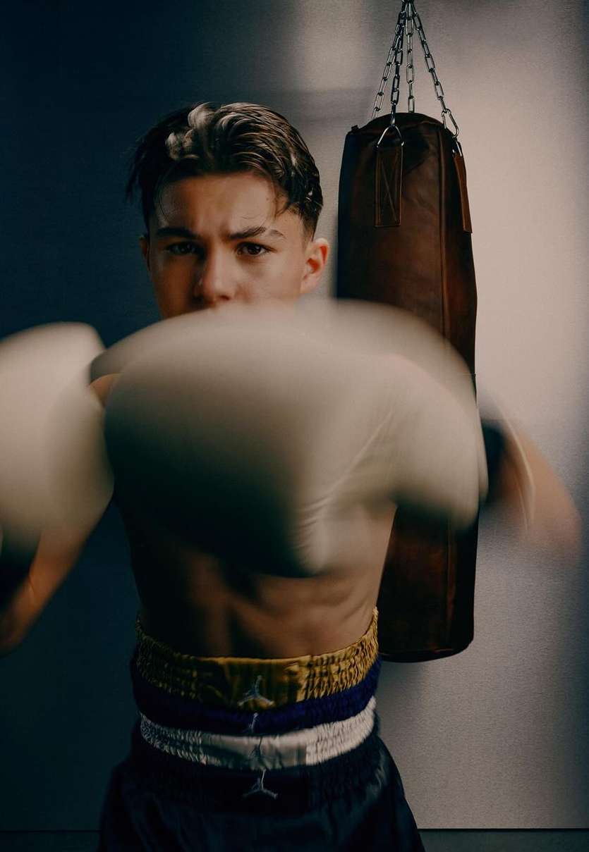 Young male boxer throwing a punch while wearing boxing gloves and athletic shorts in an indoor setting with a punching bag visible.