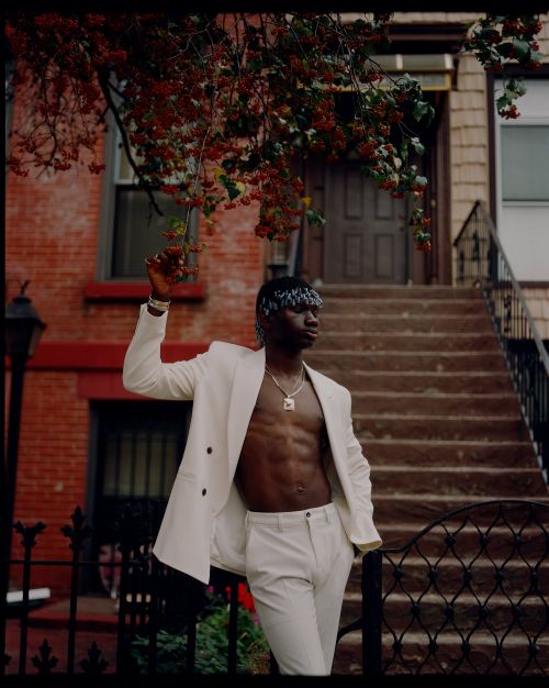 Stylish man in white suit stands in front of brick building with greenery, posing with confident expression in an urban setting.