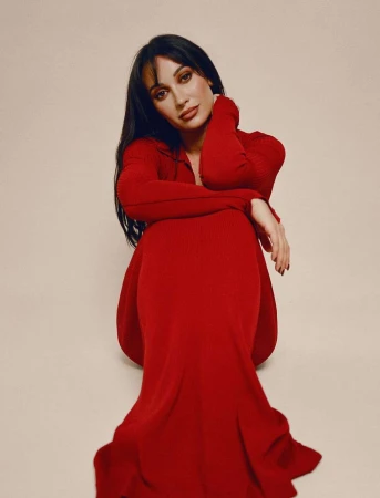 Woman with long dark hair wearing a red knit dress sitting against a plain neutral background by Kendall Bessent