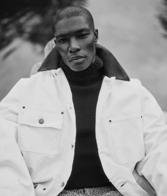 Monochrome portrait of a Black man in a dark turtleneck and off-white jacket, looking directly at the camera with a blurred background.