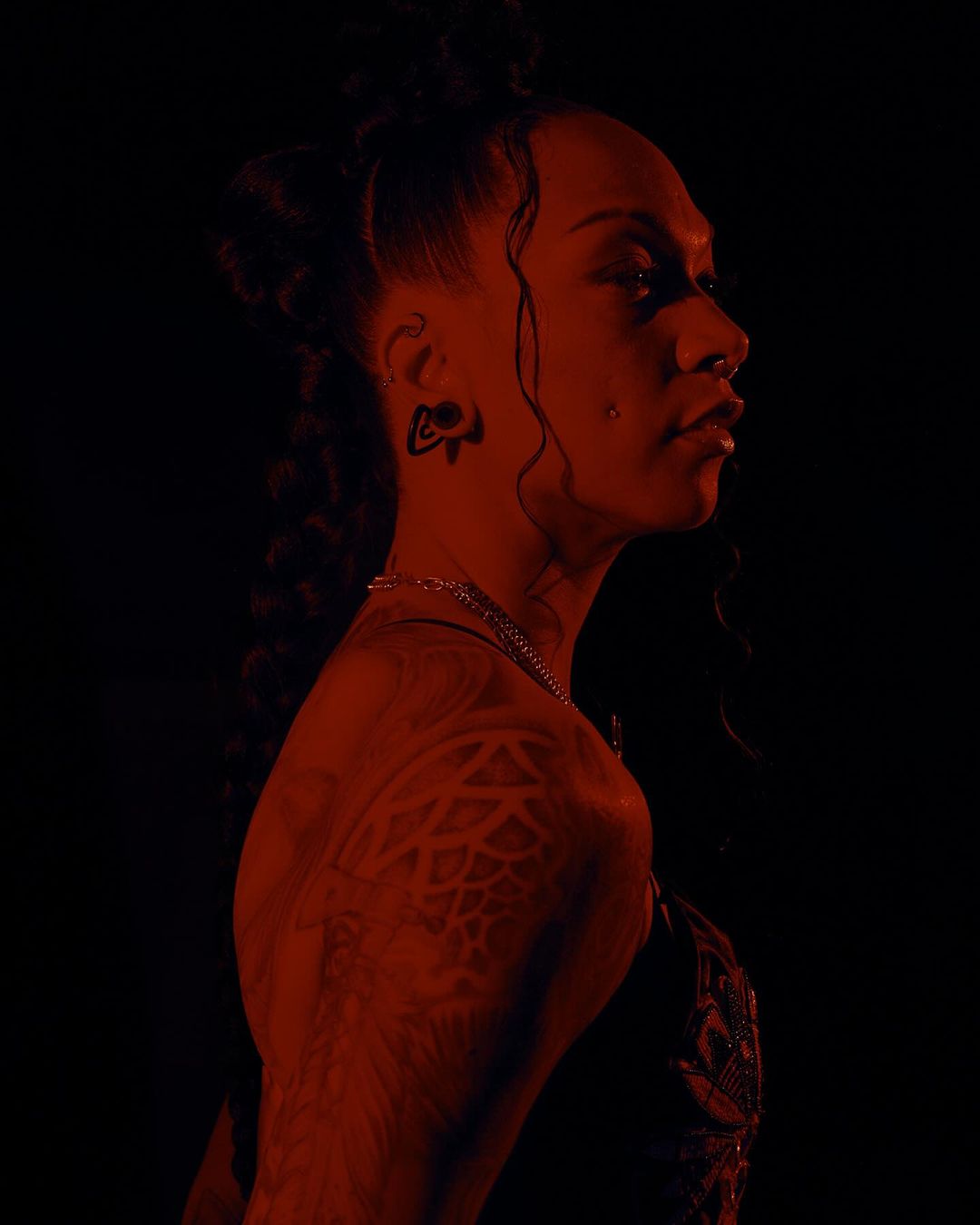 Profile portrait of a person with elaborate braids, facial piercings, and detailed tattoos lit in dramatic red light by Kendall Bessent