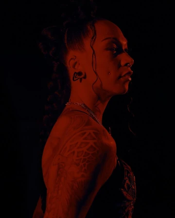 Profile portrait of a person with elaborate braids, facial piercings, and detailed tattoos lit in dramatic red light by Kendall Bessent
