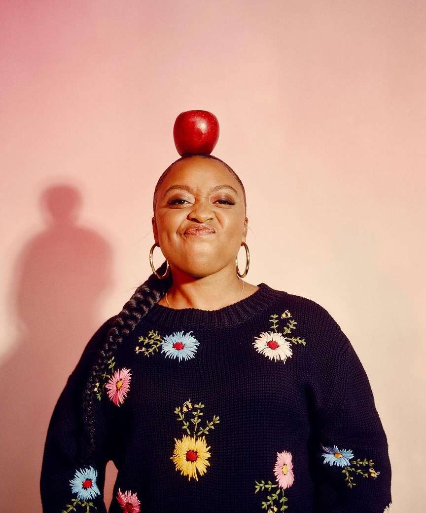 Portrait of a young Black woman with a red apple balanced on her head, making a funny face, wearing a floral embroidered sweater.