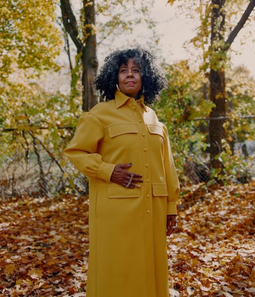 An adult female with dark hair poses in a yellow coat outdoors amid autumn leaves and trees in a full shot in natural lighting.