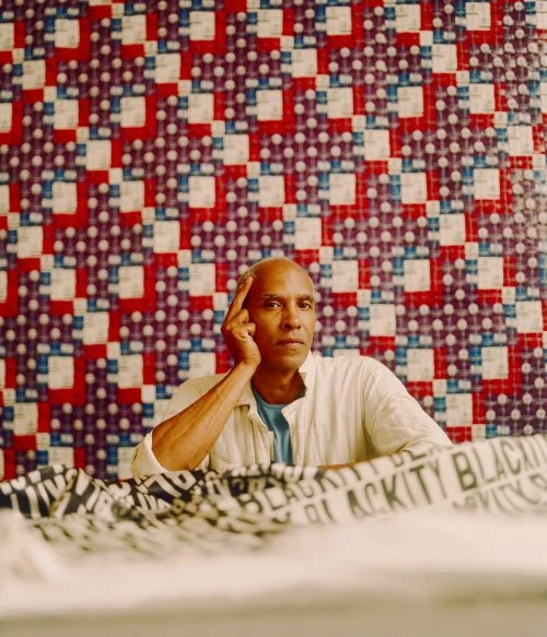 Portrait of an adult with a bald head and light skin tone. He poses indoors with a hand near his face, against a red, white, and blue patterned backdrop.