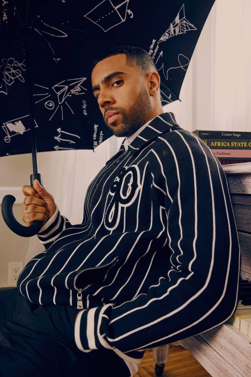 Stylish man poses indoors with umbrella; he wears a striped jacket with an embroidered emblem; books in the background. 