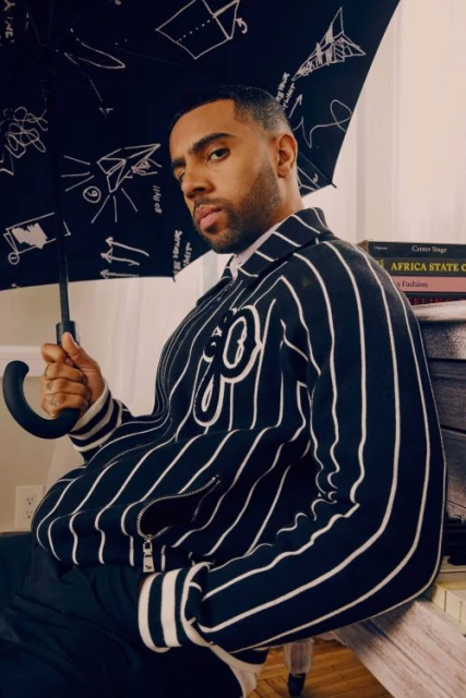 Stylish man poses indoors with umbrella; he wears a striped jacket with an embroidered emblem; books in the background. 