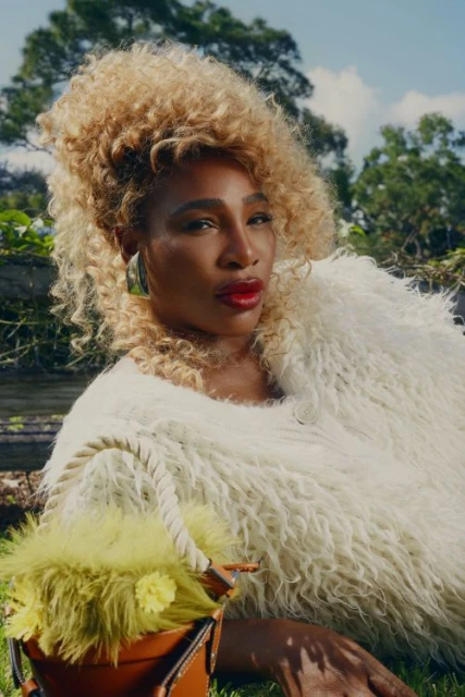 Portrait of Serena Williams with blonde Afro, wearing a white fur coat and holding a yellow bag in a green outdoor setting.