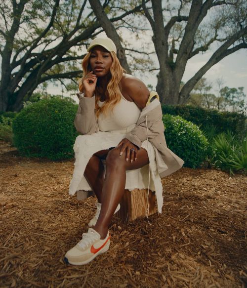 Serena Williams sits in a garden wearing an off-white dress, jacket, sneakers, and a cap in a full shot. Large trees overhead.