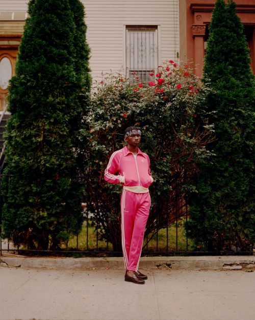An African-American man in a pink track suit stands in front of greenery and an apartment building, showcasing street style.