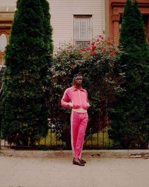 An African-American man in a pink track suit stands in front of greenery and an apartment building, showcasing street style.