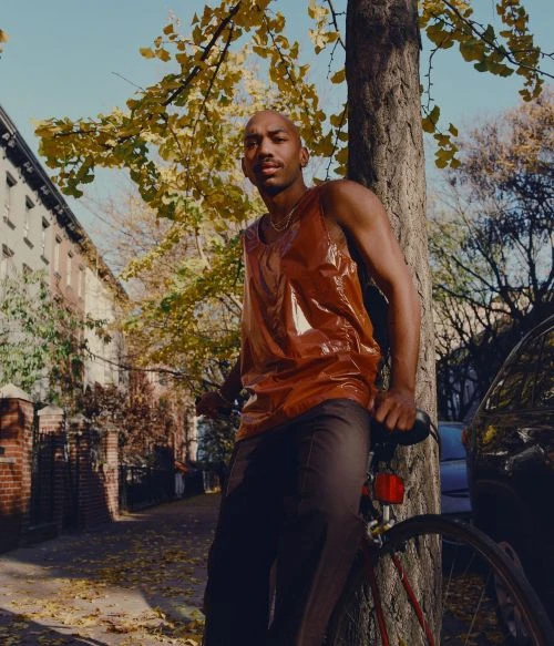 Bald man in a rust-colored tank top leans against a tree with a bicycle on a city street with fall foliage in the background.