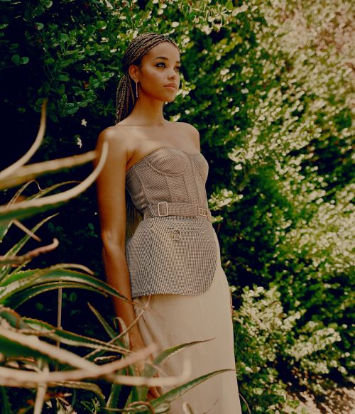 Woman with braided hair poses in strapless top and skirt amid lush greenery in a fashion-style outdoor setting, captured in soft light.