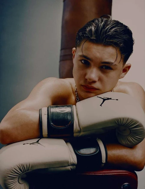 Close-up of a bare-chested young man wearing boxing gloves, leaning against a stool in front of a punching bag for fitness.