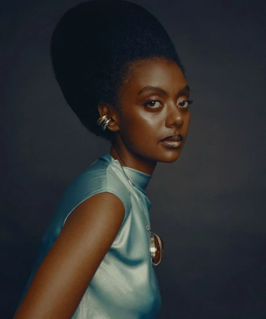 Portrait of a Black woman with a large afro wearing a teal silk top and silver jewelry, set against a solid grey backdrop in studio lighting.