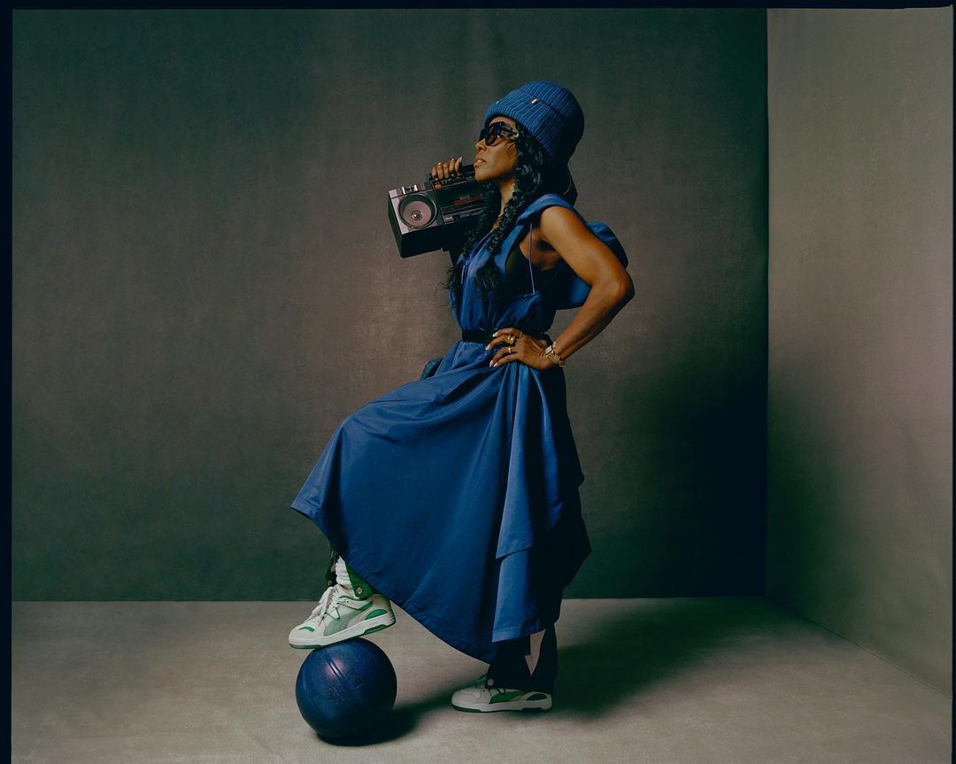 A woman in a blue dress and matching hat poses with a boombox and a basketball in a photo studio setting, side lit.