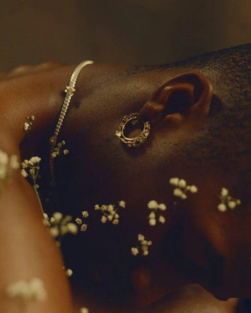 Close-up of a dark-skinned adult female with gold hoop earring and necklace, lying among small white flowers against a neutral background.