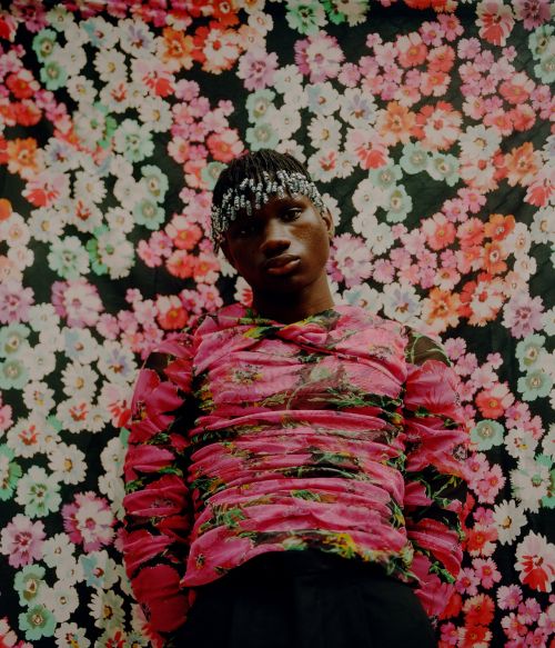 Portrait of a young adult male wearing a pink floral long-sleeve shirt, beaded headwear, and posing in front of a floral patterned backdrop.