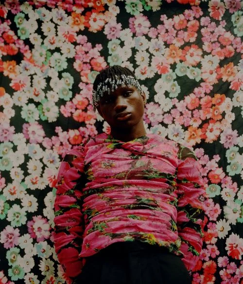 Portrait of a young adult male wearing a pink floral long-sleeve shirt, beaded headwear, and posing in front of a floral patterned backdrop.