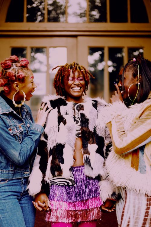 Three women stand outdoors, smiling, wearing stylish casual attire with denim, fur, and shiny fringed skirts, and large hoop earrings.