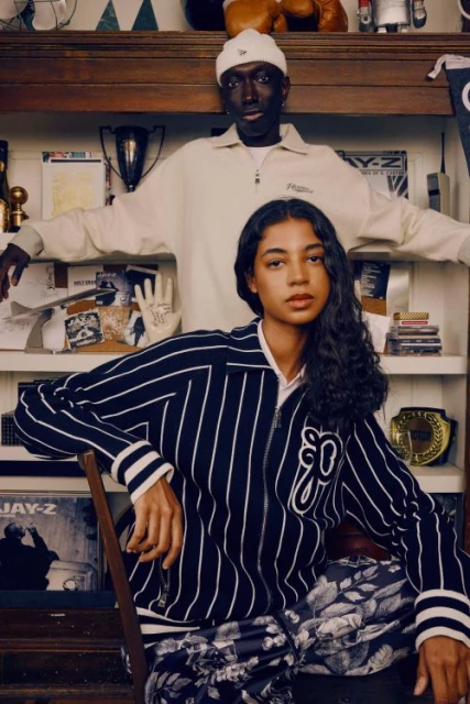 Two stylish people pose indoors in fashionable jackets and pants, with vintage decor including trophies, books, and a Jay-Z album.