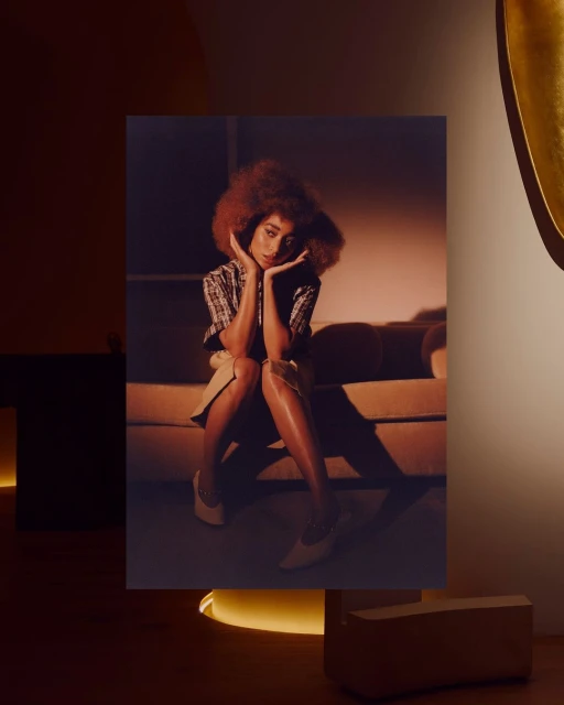 A woman with curly hair sits on a couch in a warm-toned studio setting, posing with her hands near her face and wearing heels.