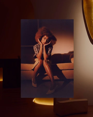 A woman with curly hair sits on a couch in a warm-toned studio setting, posing with her hands near her face and wearing heels.